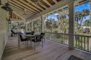 A covered outdoor patio with a wooden deck, white railings, and a table with blue chairs. - 24 Sand Dollar Drive by Avantstay Entertainers Home w/ Pool. Hot Tub, Ping Pong & Close To Beach! (Isle of Palms)