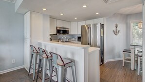 A modern kitchen with white cabinets, stainless steel appliances, and a central island with bar stools. - 103 B Port O Call by Avantstay Direct Beach Access Screened In Porch! (Isle of Palms)
