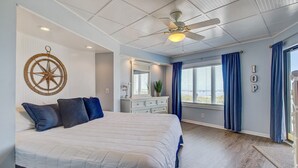 A bedroom with a large bed, a ceiling fan, a dresser, a mirror, and a window with curtains. - 103 B Port O Call by Avantstay Direct Beach Access Screened In Porch! (Isle of Palms)