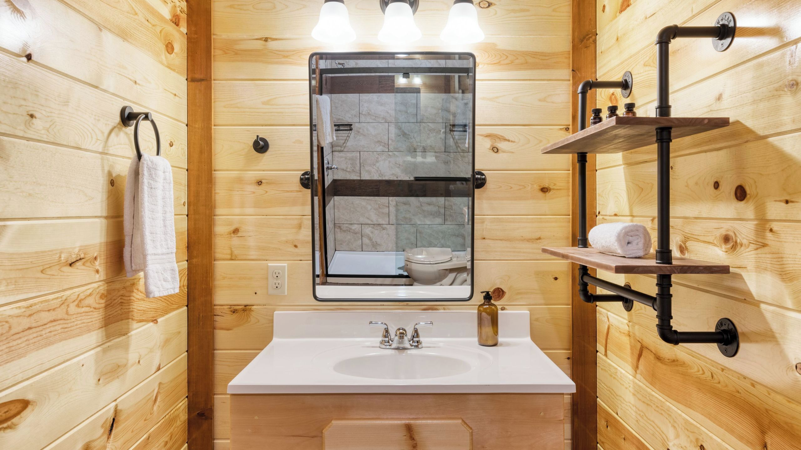 A wooden cabin bathroom with a white sink, a mirror, a glass-enclosed shower, and a shelf with toiletries.