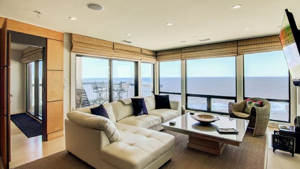 A modern living room with a large window, a sofa, a coffee table, and a wicker chair. - 1403 Ocean Club by Avantstay Beach Home w/ Great Kitchen (Isle of Palms)
