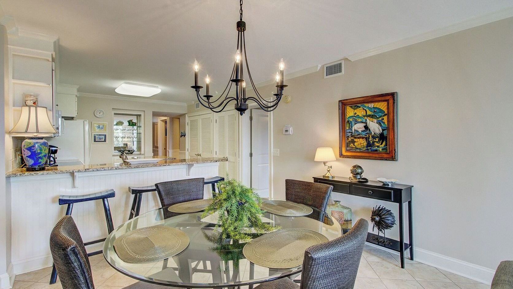 A dining area with a glass table, wicker chairs, a chandelier, a painting, and a kitchen in the background.