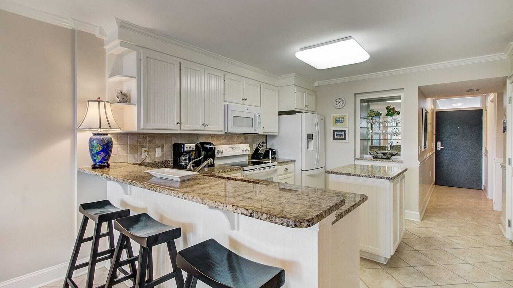 A kitchen with white cabinets, a granite countertop, a coffee maker, a microwave, and a refrigerator. There are three bar stools and a clock on the wall.