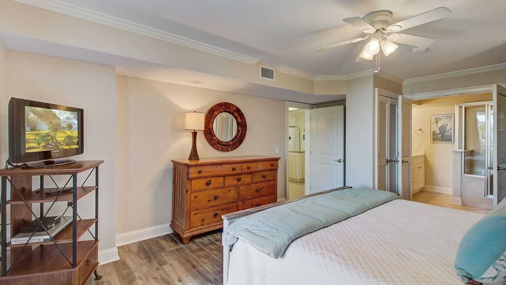 A bedroom with a bed, a dresser, a TV on a shelf, a ceiling fan, and a door leading to another room.