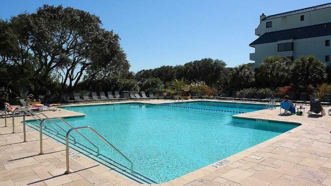 Una piscina rectangular con un borde empedrado, rodeada de sillas de descanso y un edificio al fondo.