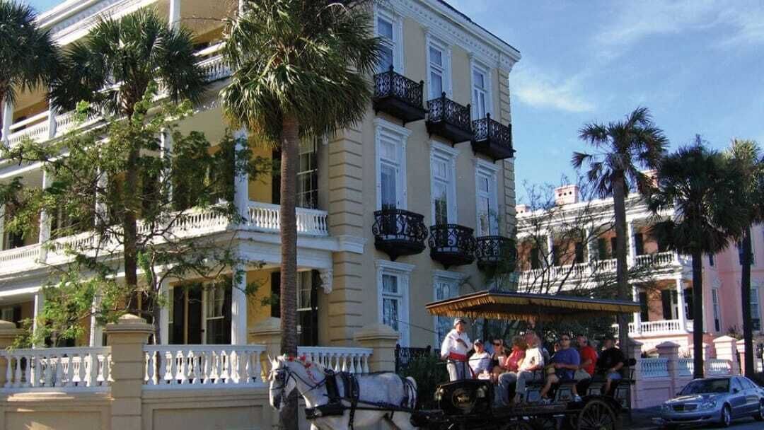 Un carruaje tirado por caballos con pasajeros, estacionado frente a un edificio de varios pisos con balcones y palmeras.