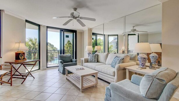 A spacious living room with a sofa, armchairs, a coffee table, and a TV. There are two lamps on side tables, and a ceiling fan. Large windows offer a view of palm trees and a building outside. - 216 C Shipwatch by Avantstay Shared Pool + Ocean Views + Walk to the Beach (Isle of Palms)