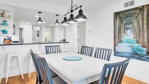 A modern kitchen with white cabinets, a black countertop, and a dining area with a white table and blue chairs. - 314 C Shipwatch by Avantstay Oceanfront With Ocean View (Isle of Palms)