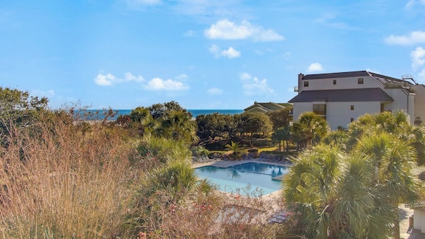 Une piscine entourée d’une végétation tropicale, un bâtiment blanc et un ciel d’un bleu limpide.