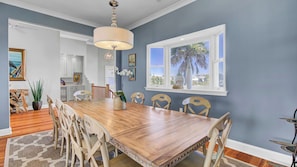 Comedor con mesa de madera, sillas color beige, una lámpara de araña y vistas a palmeras a través de una ventana.