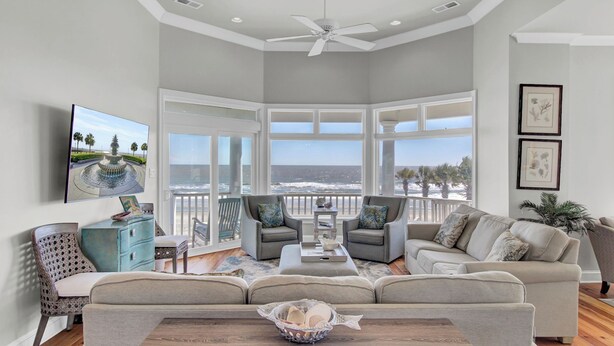 A spacious living room with a large window overlooking the ocean, a ceiling fan, and a television mounted on the wall.