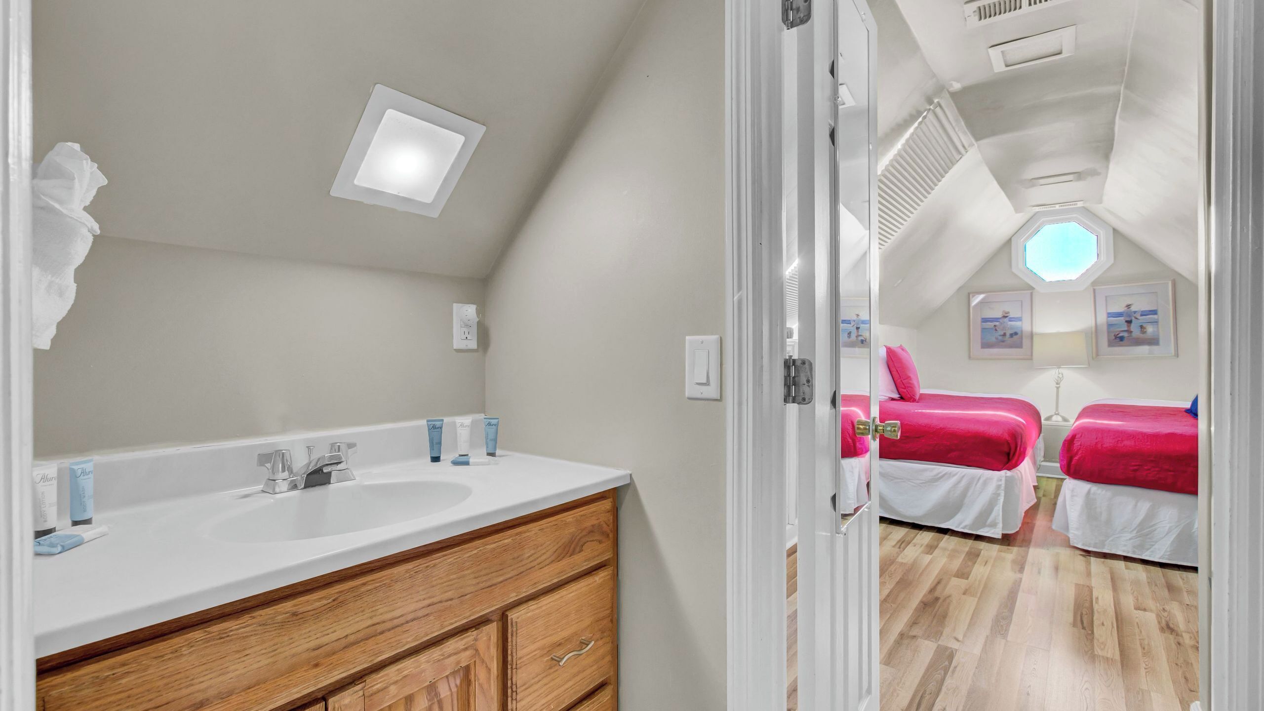 Une pièce avec une coiffeuse en bois, un lavabo blanc et un miroir. On y trouve deux lits avec une literie rouge et une fenêtre donnant sur la plage.