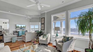 A spacious living room with a large window overlooking the ocean, a ceiling fan, and a view of the kitchen.
