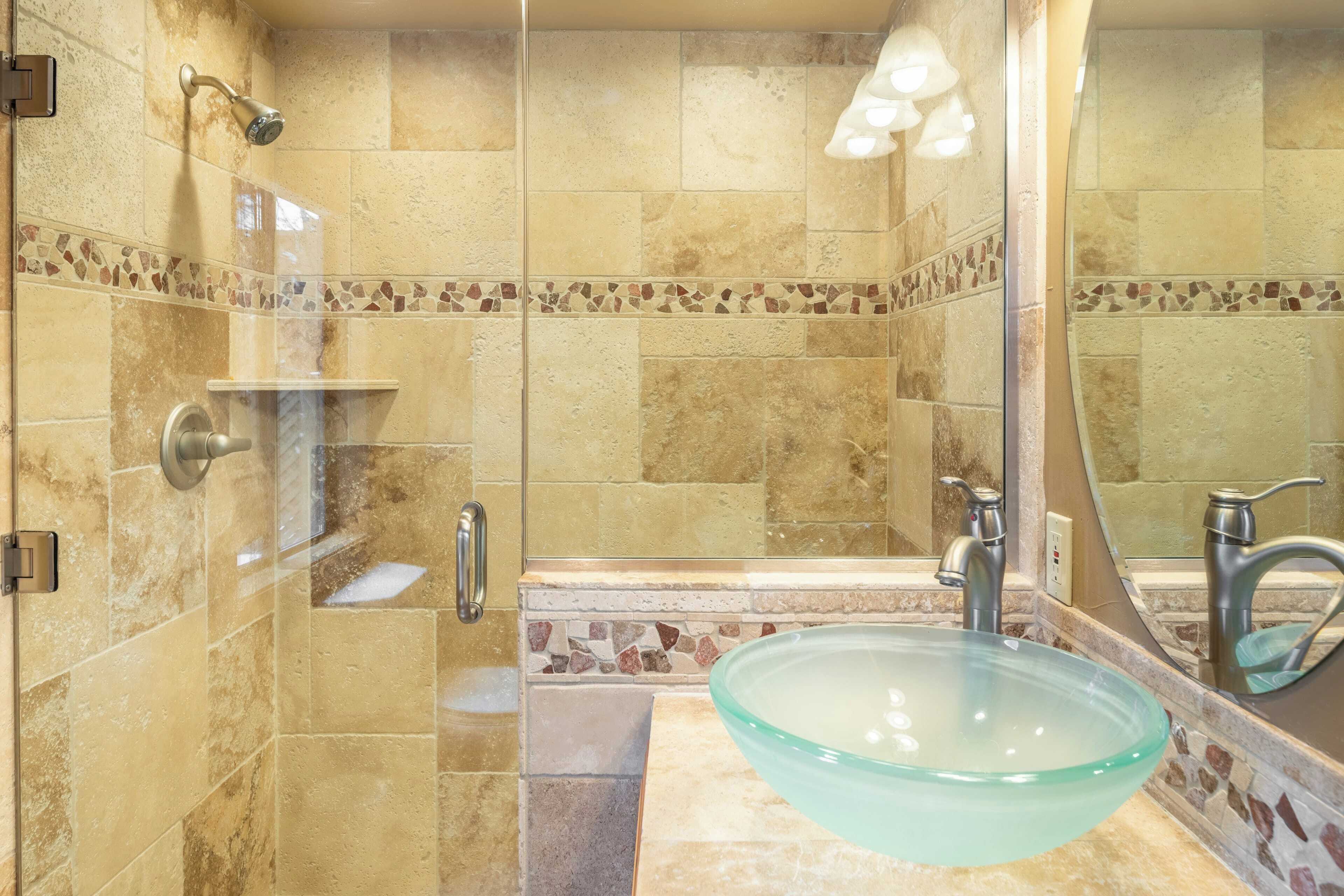 Ein Badezimmer mit einem Waschbecken aus Glas, einer Dusche mit Duschbrause und einer mit Mosaikbordüre versehenen gefliesten Wand.