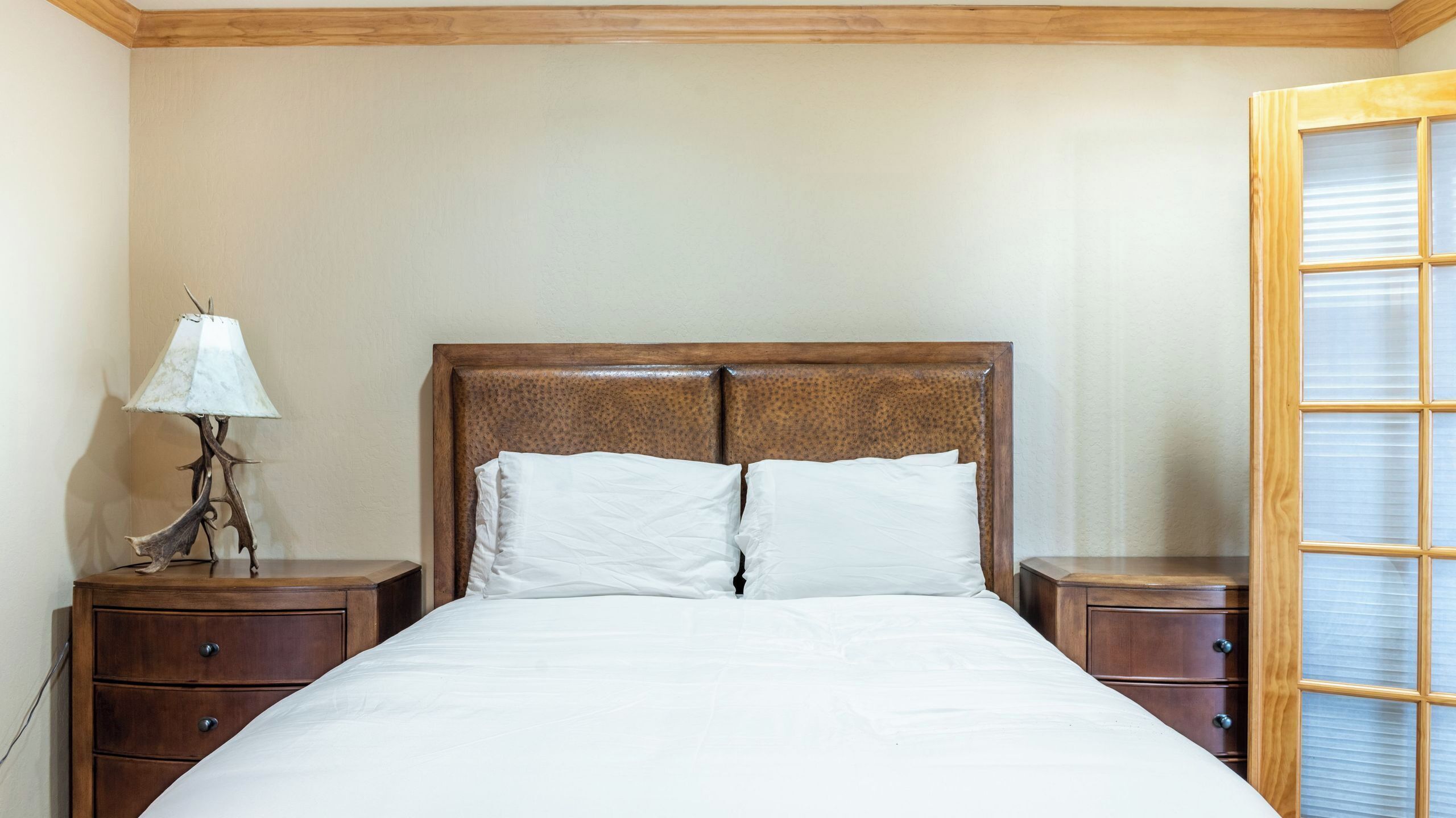 A bedroom with a wooden headboard, two white pillows, a bedside table with a lamp, and a wooden nightstand.