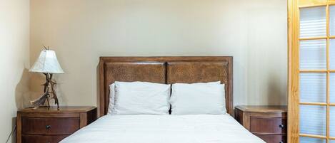 A bedroom with a wooden headboard, two white pillows, a bedside table with a lamp, and a wooden nightstand.