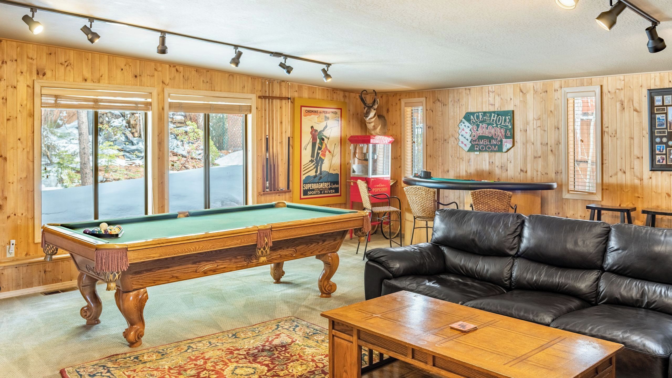 A pool table, a leather couch, and a billiards table in a room with wooden walls and windows.