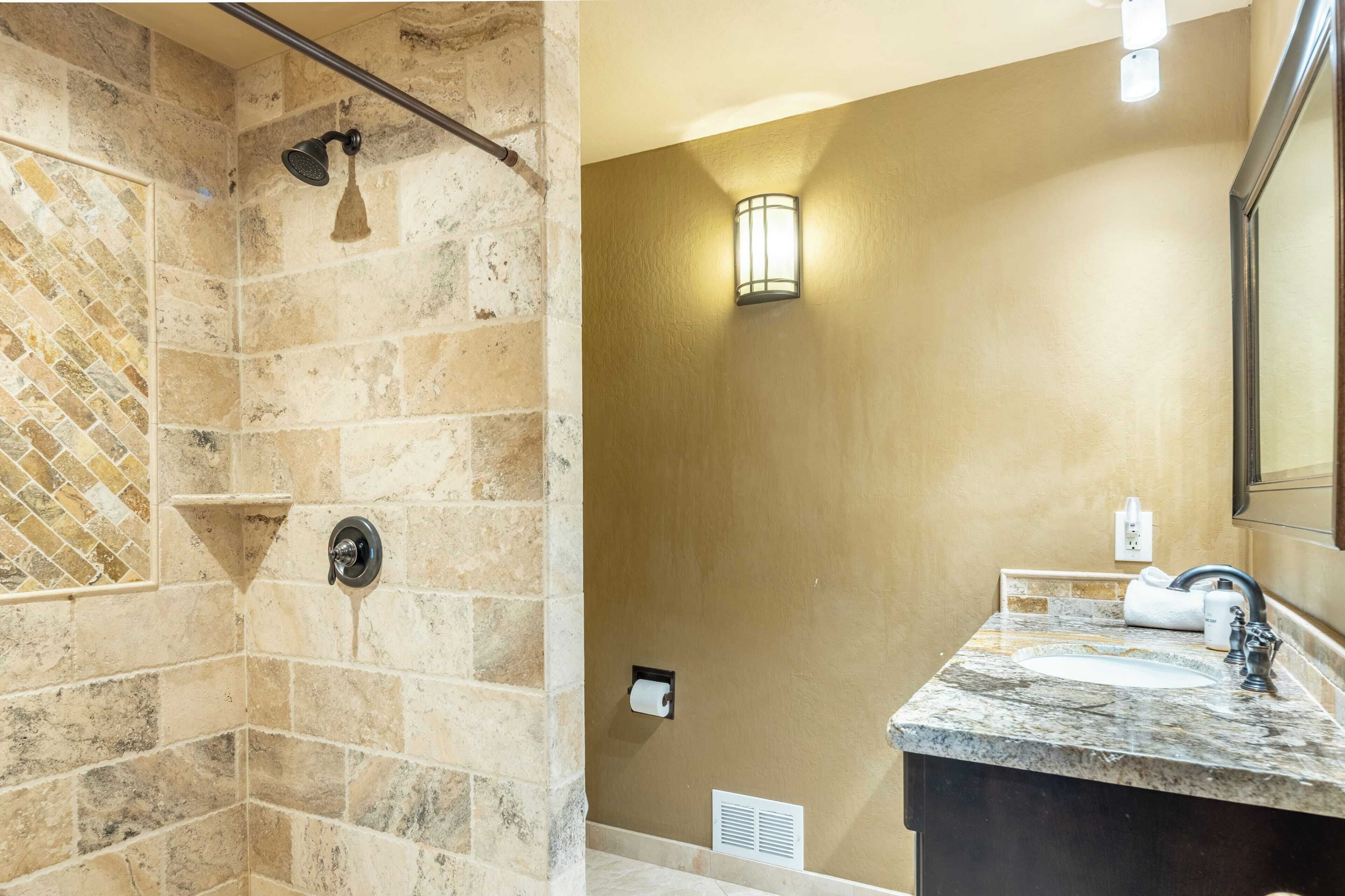 A bathroom with a shower area featuring stone tiles and a handheld showerhead, a sink with a marble countertop, and a wall-mounted light fixture.