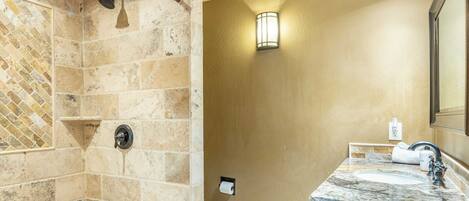 A bathroom with a shower area featuring stone tiles and a handheld showerhead, a sink with a marble countertop, and a wall-mounted light fixture.