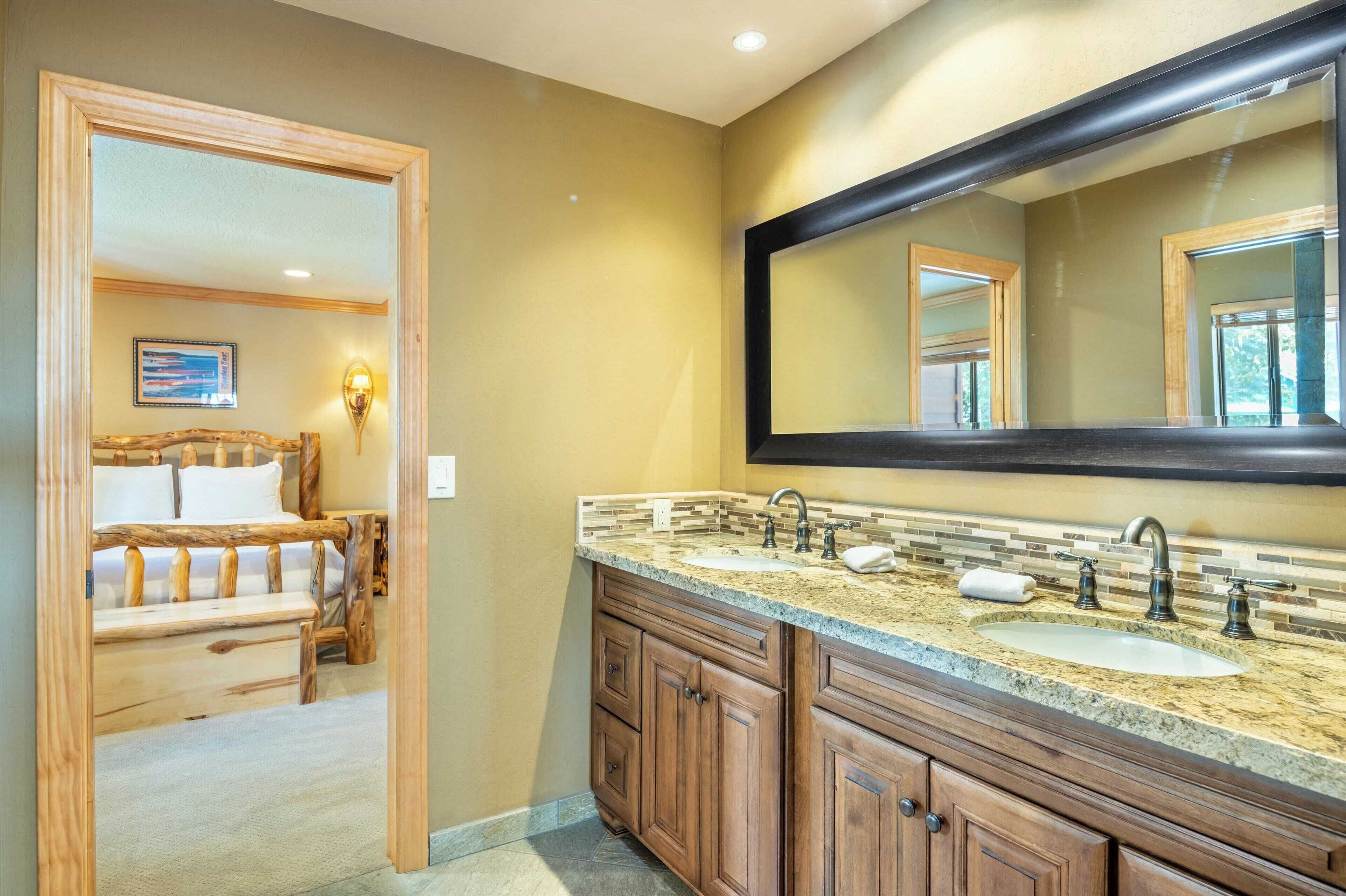 Une salle de bain avec un grand miroir, des meubles en bois, un lavabo et un lit visible à travers une porte ouverte.
