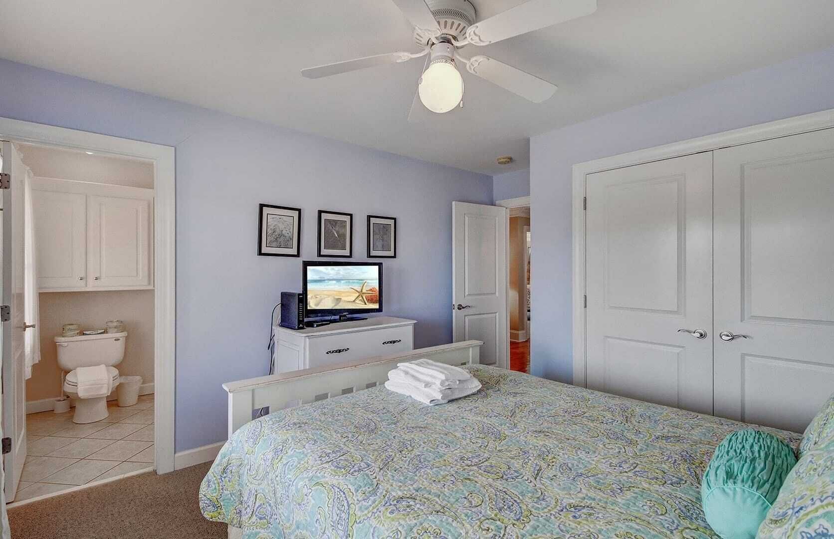 A bedroom with a bed, a television, a ceiling fan, and a bathroom with a toilet and cabinets.