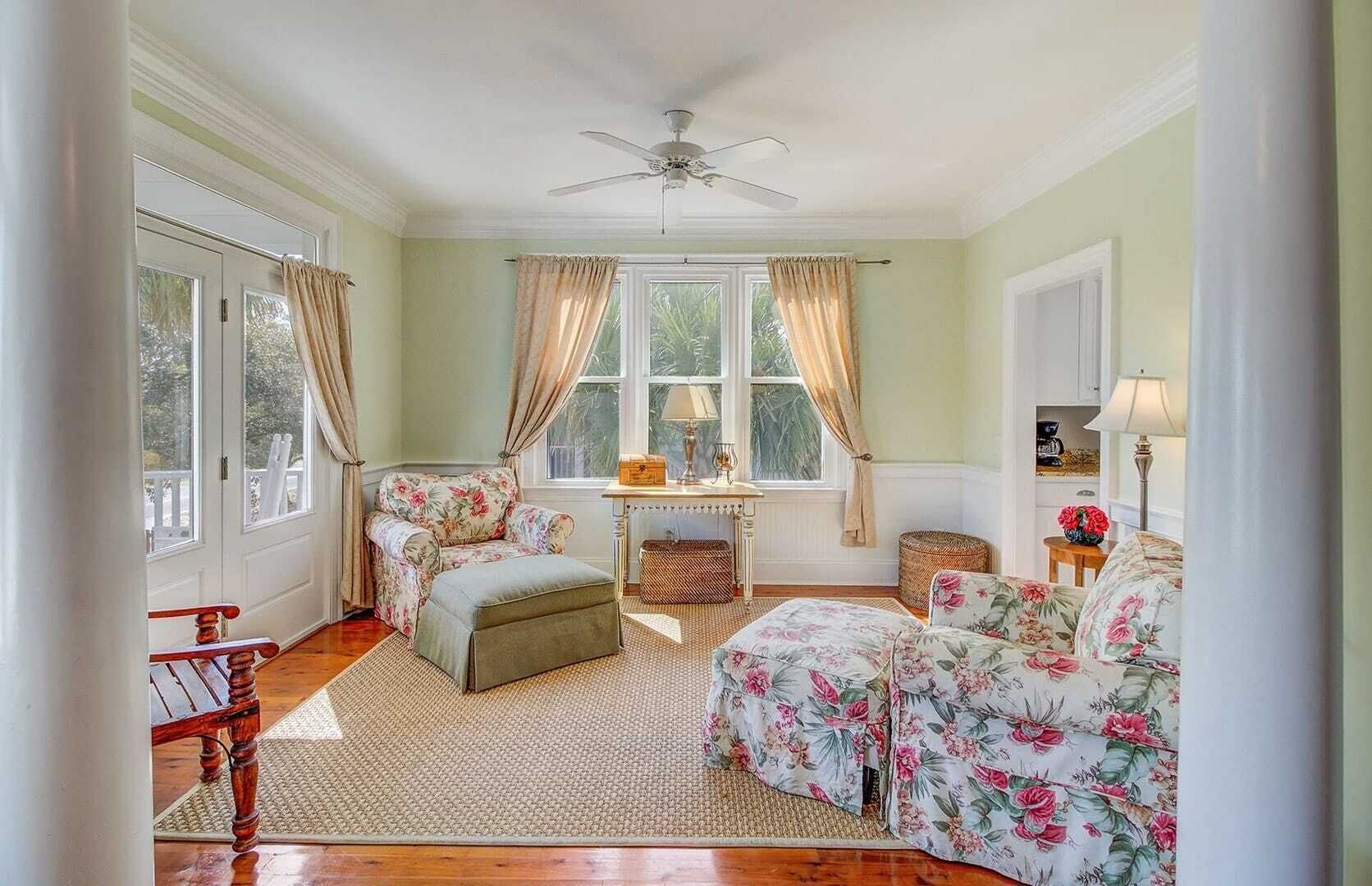 Ein Zimmer mit Holzboden, Deckenventilator, Fenster mit Vorhängen, einem kleinen Tisch mit Lampe und floral gepolsterten Möbeln.
