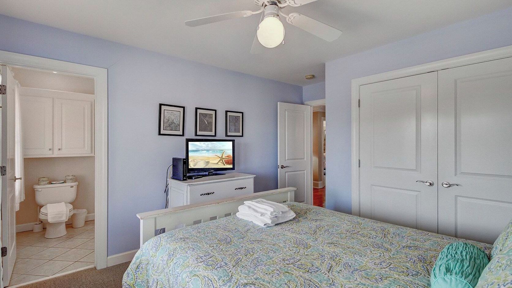 Een slaapkamer met een bed, een televisie, een plafondventilator en een deur naar een badkamer.