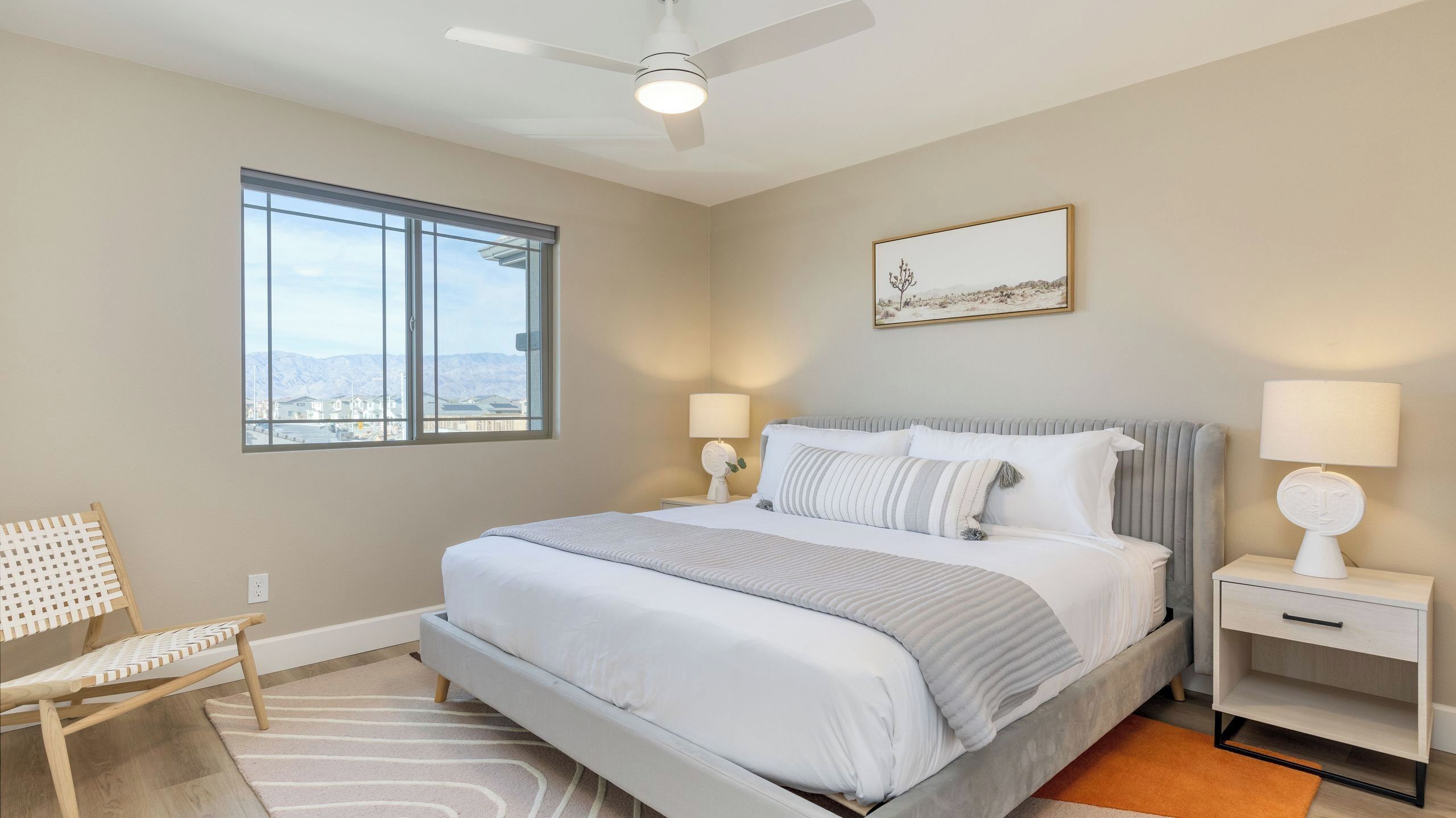 Une chambre à coucher comprenant un lit, deux tables de chevet, une chaise, un ventilateur de plafond, une fenêtre avec vue et un tableau encadré au mur.