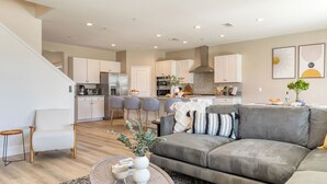 A modern living room with a grey sofa, a coffee table with decorative items, a kitchen in the background with white cabinets and a stainless steel refrigerator. - Sleeps 16! Pool Oasis w/ Game Room, Hot Tub, Fire Pit Desert Eden by Avantstay (Coachella)
