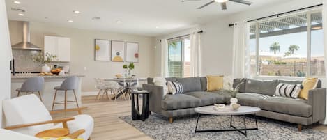 A modern living room with a gray sofa, white armchairs, and a coffee table. There is a kitchen in the background with white cabinets and a dining area with a table and chairs.