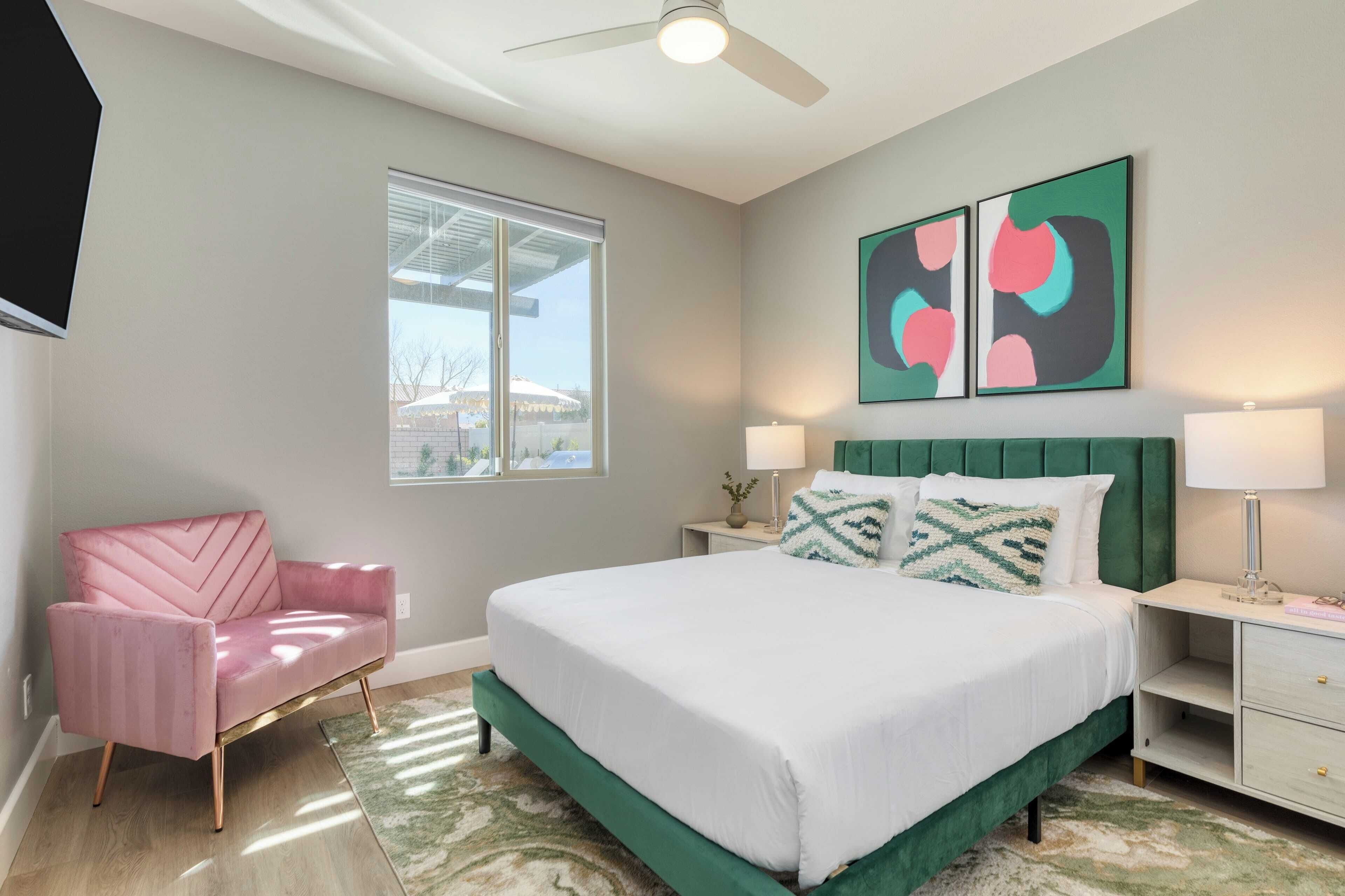 A modern bedroom with a green upholstered bed, a pink chair, and artwork on the wall.