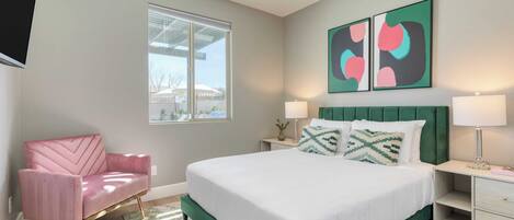A modern bedroom with a green upholstered bed, a pink chair, and artwork on the wall.
