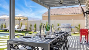 An outdoor patio with a dining table set for six, surrounded by chairs. There is a grill and a red cart in the background. - Sleeps 16! Pool Oasis w/ Game Room, Hot Tub, Fire Pit Desert Eden by Avantstay (Coachella)