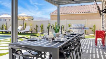 Un patio al aire libre con una mesa de comedor preparada para seis personas, rodeada de sillas. Se ve una parrilla y un carrito rojo al fondo.