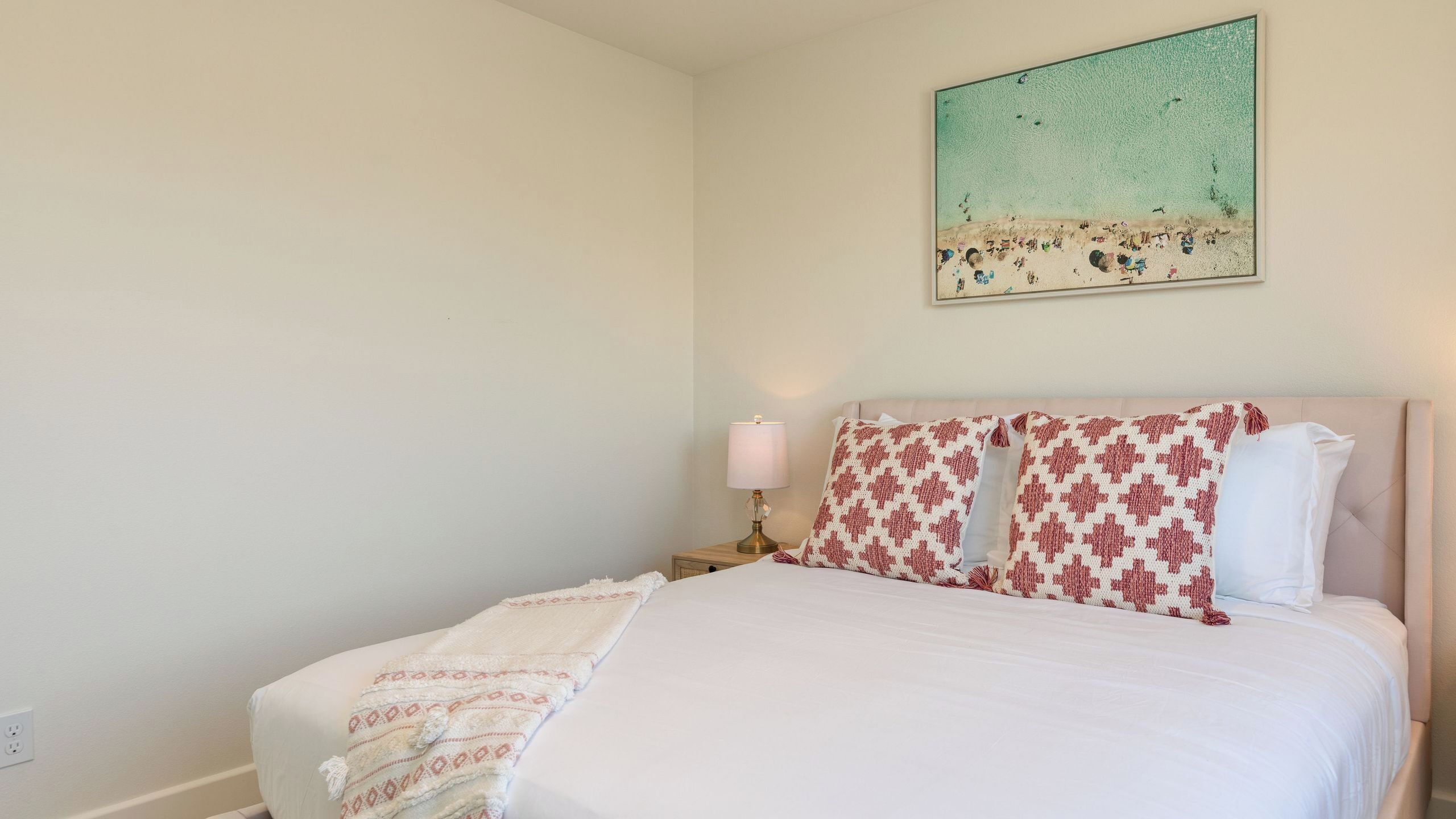 Een slaapkamer met een bed, een nachtkastje met een lamp, een wanddecoratie van een strandgezicht en een stopcontact op de muur.