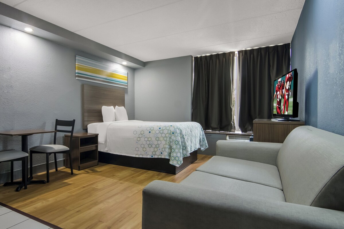 Habitación de hotel con cama, sofá, escritorio con sillas, televisor y ventana con cortinas.