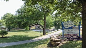 A park sign for North Aurora Riverfront Park with a pathway, trees, and a small building in the background. - HomeTowne Studios by Red Roof & Conference Center Chicago – N Aurora/ Naperville (North Aurora)