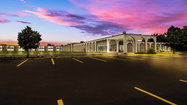 A parking lot with a building featuring arched windows and a flat roof. - HomeTowne Studios by Red Roof & Conference Center Chicago – N Aurora/ Naperville (North Aurora)