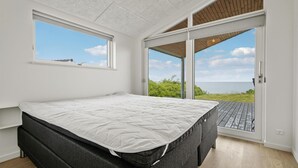 A bedroom with a large bed, a window, and a sliding glass door leading to an outdoor area with a view of the sea.