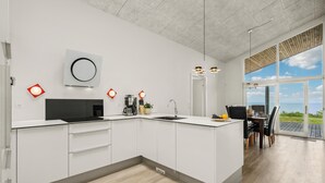 A modern kitchen with white cabinetry, a black countertop, and a large island. There are pendant lights above the island and a view of the outdoors through large windows.