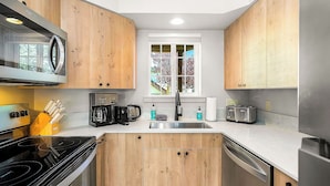A modern kitchen with wooden cabinets, stainless steel appliances, a window with a view, and a countertop with various items. - Icicle Village Blizzard Pool Parking (Leavenworth)