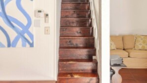 A wooden staircase leading to a second floor, a white wall with a blue abstract design, and a beige sofa in the background.