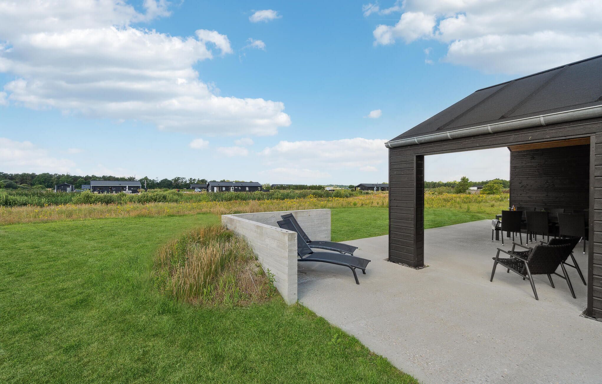Un área de estar moderna al aire libre con sillones reclinables y una mesa, un porche cubierto y vistas a un campo y edificios a lo lejos.