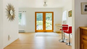A spacious room with a wooden floor, a large window with a view of greenery, and a dining area with red chairs. - Ski - Luxury Retreat Home - Stream, Waterfalls! (Palenville)