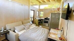 Ein Schlafzimmer mit einem Bett, einem kleinen Tisch mit Gegenständen, einer Küchenzeile mit Kühlschrank und einem an der Wand befestigten Fernseher.