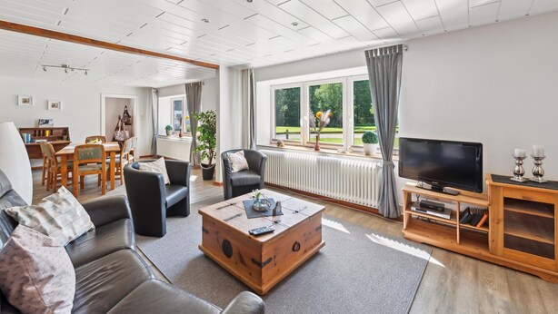 A spacious living room with a sofa, coffee table, and TV. There is a dining area with a table and chairs, and a view of greenery outside.