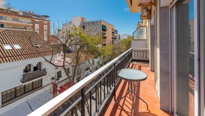Un balcon avec une table et des chaises, donnant sur une rue bordée d’immeubles et d’arbres.