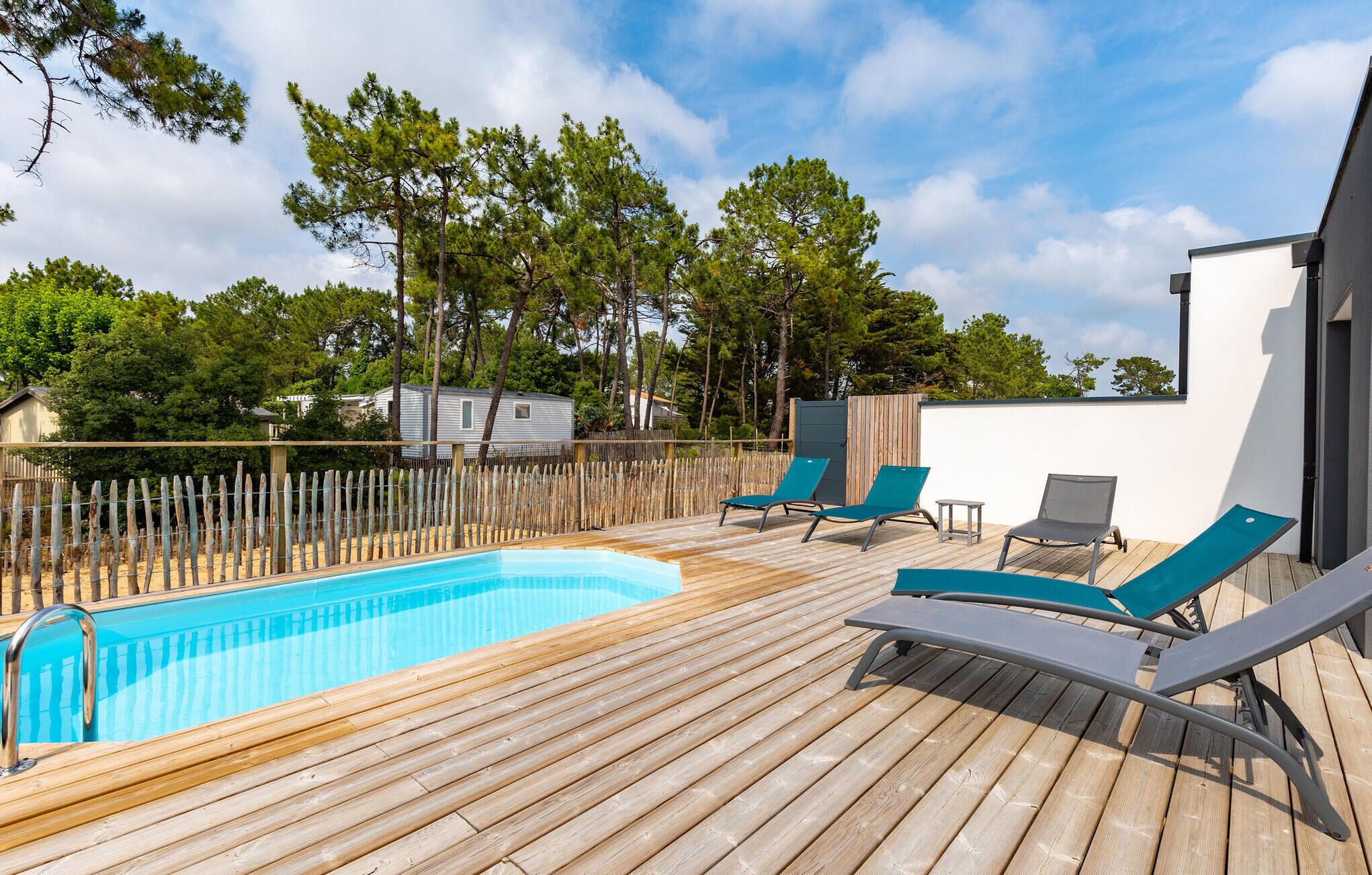 A wooden deck with a swimming pool, lounge chairs, and a fenced area with trees.