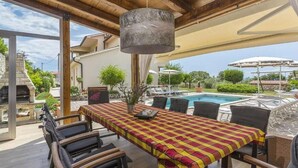 An outdoor dining area with a checkered tablecloth, wooden chairs, a hanging light fixture, and a view of a swimming pool and greenery. - Beautiful home in Koromacno-Brovinje (Koromacno-Brovinje)