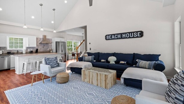 A spacious living room with a large sofa, a coffee table, and a dining area with bar stools. A sign on the wall reads "BEACH HOUSE". - Cape Cod: Gimlet Getaway (Eastham)